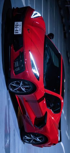 a red sports car hanging from the side of a wall with its hood up and lights on