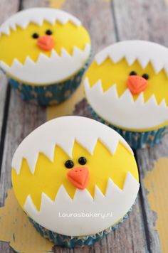 three decorated cupcakes with yellow and white frosting on wooden boards, one has an egg in the shape of a chicken