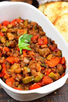 a white casserole dish filled with meat and veggies next to bread