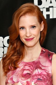 a woman with long red hair wearing a pink dress and smiling at the camera while standing in front of a black background