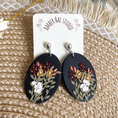 earrings with flowers painted on them sitting on a woven tablecloth next to a card