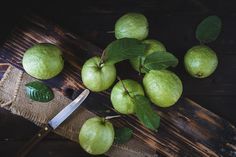 some green apples are sitting on a wooden table with a knife and leafy leaves