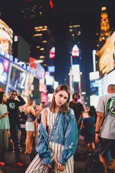 a woman standing in the middle of a busy city street at night wearing a denim jacket and striped pants