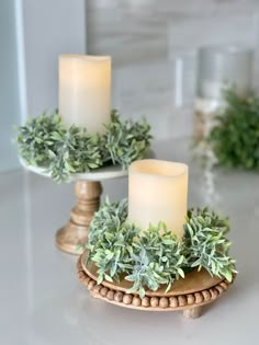 two white candles sitting on top of a table next to each other with greenery around them