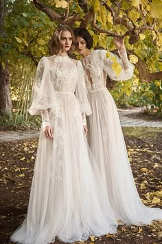 two women standing next to each other under a tree in white dresses with long sleeves