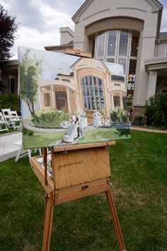 an easel with a painting on it in front of a large house and lawn