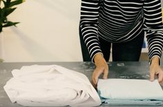 a woman is rolling up some white sheets on a table with a potted plant in the background