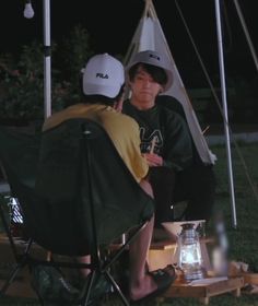 two people sitting in lawn chairs at night with one person wearing a hat and the other holding an umbrella