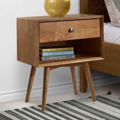 a nightstand with books and a yellow vase on it next to a bed in a bedroom