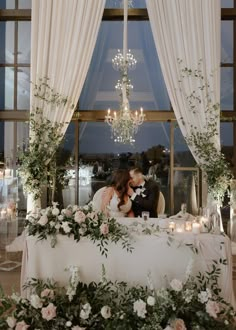 a bride and groom kissing under a chandelier at their wedding reception in front of a window