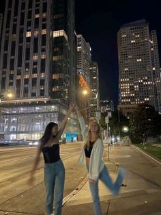 two women are dancing on the sidewalk in front of some tall buildings at night time