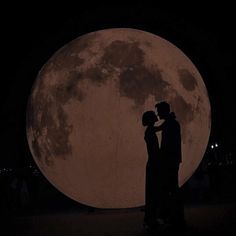 two people standing in front of a large full moon at night with their arms around each other