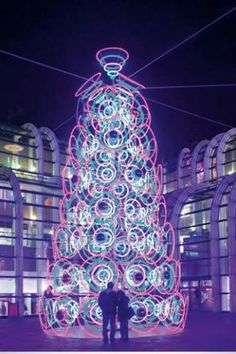 two people standing in front of a lit up christmas tree at night with lights on it