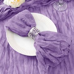 a white plate topped with purple napkins next to a wine glass and flower vase