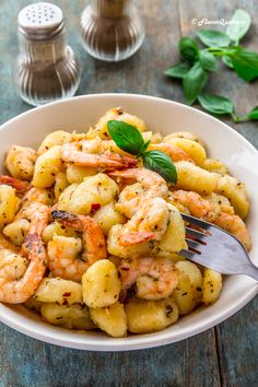 a white bowl filled with pasta and shrimp on top of a wooden table next to two salt shakers