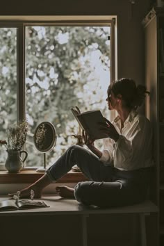 a woman sitting on a window sill reading a book in front of a window