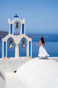 a woman in a white dress is walking near bells on a building overlooking the water