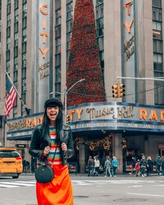 a woman in an orange skirt and black jacket is standing on the street near a christmas tree