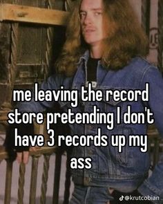 a man with long hair wearing a jean jacket and jeans standing in front of a wooden fence