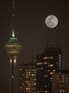 a full moon is seen over the top of a tall building with lights on it