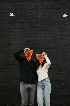 two people standing in front of a black wall holding up slices of pizza to their faces