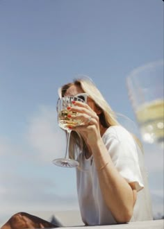 a woman sitting on the ground holding a wine glass up to her face and looking into the distance