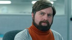 a man with a beard and an orange shirt is looking at the camera while sitting in front of a computer desk