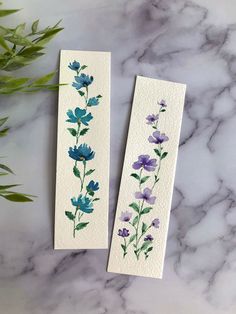 two bookmarks with watercolor flowers on them sitting on a marble surface next to a plant