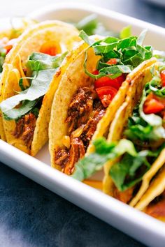 three tacos with meat, lettuce and tomatoes on a white plate sitting on a table