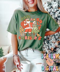 a woman wearing a santa clause t - shirt next to a christmas tree and holding a coffee mug