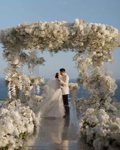 a bride and groom standing under an archway with white flowers in front of the ocean