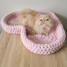 a cat sitting in a pink crocheted bed on top of a wooden table