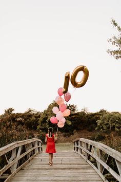 a woman in a red dress holding pink and gold balloons while standing on a bridge