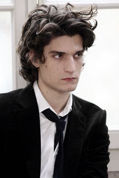 a young man with curly hair wearing a suit and tie, looking at the camera