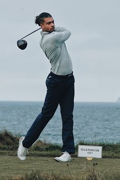 a man swinging a golf club on top of a grass covered field next to the ocean