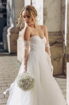 a woman in a white wedding dress holding a bridal bouquet and looking down at the ground