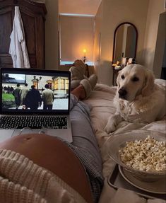 a dog laying on a bed next to a laptop and bowl of popcorn with people in the background