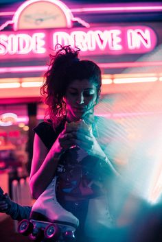 a woman standing in front of a neon sign holding a skateboard and looking at her phone