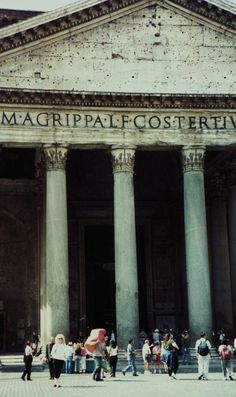 many people are walking around in front of a large building with columns and letters on it