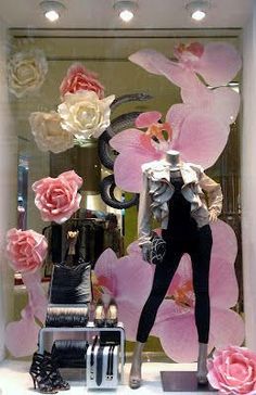 a window display with pink flowers and mannequins in the front, behind which is an image of a woman's dress