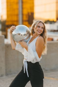 a woman holding a silver ball in her right hand and smiling at the camera with both hands behind her back