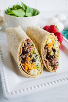 two burritos on a plate with strawberries and spinach in the background