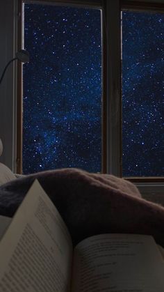 an open book sitting on top of a bed next to two windows with stars in the sky