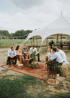 a group of people sitting on top of a rug under a white tent next to a field