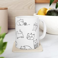 a white coffee mug with black cats on it sitting next to some lemons and an avocado