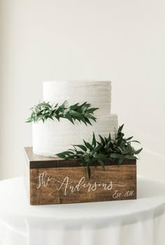 two tiered wedding cake with greenery on top, sitting on a wooden box