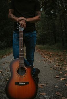 a man holding an acoustic guitar in the middle of a road with leaves on the ground