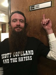 a man with a long beard is standing in front of a door and pointing at the camera