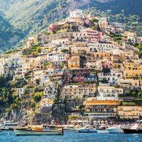 boats in the water near a hillside with houses on it's sides and mountains behind them