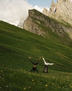 two people are standing in the middle of a grassy field with mountains in the background